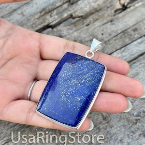 May include: A rectangular pendant featuring a deep blue lapis lazuli stone with natural gold-colored pyrite flecks, encased in a polished silver-toned metal frame with a simple triangular bail at the top for attachment to a chain or cord.