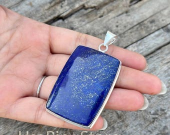 May include: A rectangular pendant featuring a deep blue lapis lazuli stone with natural gold-colored pyrite flecks, encased in a polished silver-toned metal frame with a simple triangular bail at the top for attachment to a chain or cord.