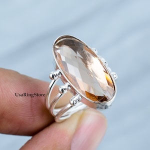 Peut inclure: Une bague en argent avec une grande pierre précieuse ovale de couleur brun clair. La bague a un design délicat à trois bandes avec de petites perles argentées.