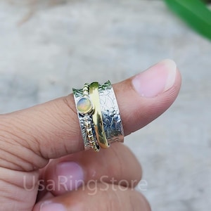 Puede incluir: Un anillo giratorio de plata y oro con un diseño floral y una pequeña piedra de ópalo. El anillo se lleva en el pulgar.
