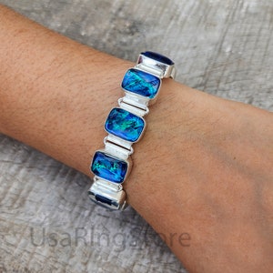 May include: A silver bracelet with five rectangular blue and green opal stones. The bracelet is on a person's wrist.