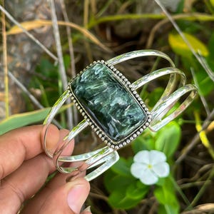 May include: A silver cuff bracelet featuring a large green and grey gemstone set in a silver bezel. The bracelet has three bands and is decorated with small silver beads.