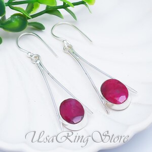 May include: A pair of silver dangle earrings with oval, faceted, ruby-red gemstones. The earrings have a modern design with a long, slender silver bar and a hook closure. The text "Usa Ring Store" is visible.
