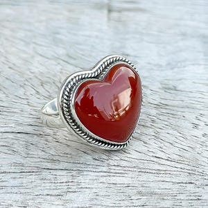 May include: A silver ring featuring a red heart-shaped carnelian gemstone. The ring has a detailed, rope-like design around the edge of the stone.