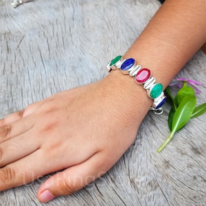 May include: A silver bracelet featuring a series of oval-shaped gemstones in green, blue, and red. The bracelet is worn on a wrist, showcasing the colorful stones against the skin. The bracelet is on a wooden surface.