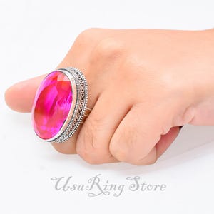 Pode incluir: Um anel de prata com uma grande gema rosa de forma oval. O anel tem um design decorativo e ornamentado em torno da gema.