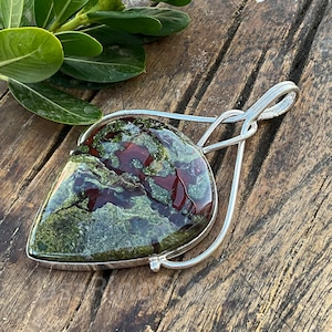 May include: A teardrop-shaped green, red, and black stone pendant with a silver setting. The pendant is on a wooden surface.