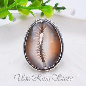 Può includere: Anello ovale in argento con una conchiglia di ciprea naturale con un motivo marrone e bianco. Il guscio è incastonato in una fascia d'argento, creando un design unico e organico. Un gioiello di tendenza.
