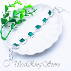 May include: A silver-toned chain bracelet featuring six square-cut emerald green stones set in metal bezels. The bracelet includes a circular clasp and a series of round links for adjustment. The text "UsaRing Store" is printed at the bottom.
