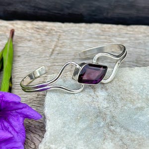 Puede incluir: Un brazalete de plata con un cierre en forma de corazón y una piedra preciosa púrpura en el centro. El brazalete tiene un diseño delicado y fluido.