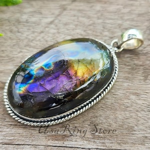 May include: An oval pendant featuring an iridescent gemstone with purple, blue, and yellow hues. The stone is set in a silver-toned metal frame with a rope design. A silver bail is at the top for a chain. The pendant is a statement piece.