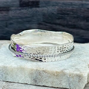 Puede incluir: Un brazalete de plata martillado con una banda texturizada y un detalle de alambre con cuentas.