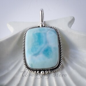 May include: A silver pendant featuring a light blue and white gemstone. The gemstone is set in a silver frame with a rope-like design. The pendant is displayed against a white background.
