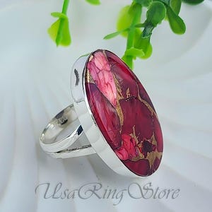May include: A silver ring with a large, oval-shaped stone. The stone is a deep red color with gold veining. The ring has a polished finish and a simple band. The text "Usa Ring Store" is visible.