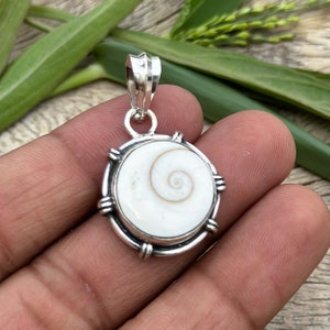 Puede incluir: Un colgante de plata con una concha blanca con un patrón en espiral. El colgante es redondo y tiene un borde de plata con acentos decorativos.
