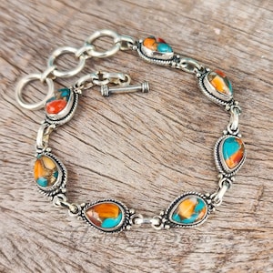 Könnte beinhalten: Ein silbernes Armband mit tropfenförmigen Steinen in Türkis, Orange und Rot. Die Steine sind in Silberfassungen gefasst und durch eine Silberkette mit einem Knebelverschluss verbunden. Das Armband liegt auf einer Holzoberfläche. Die Länge beträgt ca. 19 cm.
