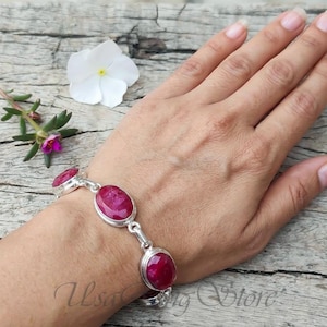 Puede incluir: Una pulsera de plata con piedras preciosas ovaladas de color rojo intenso. La pulsera se lleva en la muñeca, con las piedras engastadas en biseles individuales de plata y unidas entre sí. El fondo incluye una flor blanca y una superficie de madera.