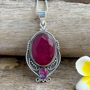 Op de afbeelding: Een zilveren hangerketting met een grote, ovaal geslepen robijn. De robijn heeft een dieprode kleur en is gezet in een decoratief zilveren montuur. Onder de robijn bevindt zich een kleinere, iriserende edelsteen. De ketting wordt gepresenteerd op een houten oppervlak.