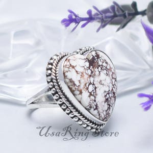 Może przedstawiać: Srebrny pierścionek z kamieniem w kształcie serca w kolorze białym i brązowym. Kamień otoczony jest koralikową obwódką. Pierścionek leży na białej powierzchni, z fioletowymi kwiatami w tle. Widoczny jest tekst "UsaRing store".