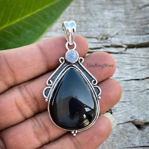 May include: A teardrop-shaped black onyx pendant set in a polished silver frame with decorative scrollwork. A small, round, iridescent moonstone is positioned above the main stone, leading to a silver bail. The text UsaRingStore is visible.