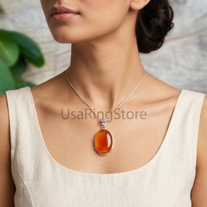 May include: A silver necklace featuring an oval-shaped, translucent orange carnelian gemstone pendant set in a smooth silver bezel. The pendant hangs from a delicate silver chain. The text UsaRingStore is visible across the centre of the image.