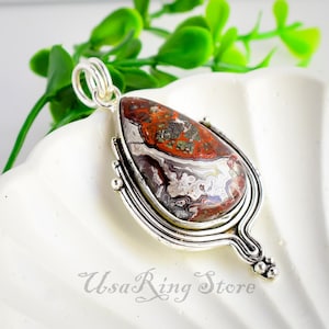 Puede incluir: Un colgante de plata con una piedra de ágata roja, blanca y gris en forma de lágrima. El colgante tiene un diseño de plata detallado alrededor de la piedra.