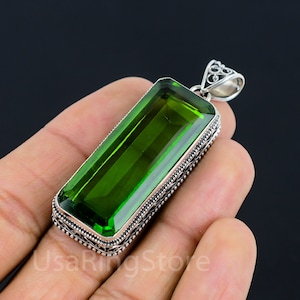 Puede incluir: Un colgante de plata con una gran piedra rectangular de color verde esmeralda. El colgante tiene un diseño detallado y adornado alrededor de la piedra preciosa.