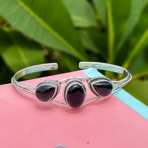 Puede incluir: Un brazalete de plata con tres acentos de piedras preciosas negras. El brazalete está abierto y tiene un diseño delicado.