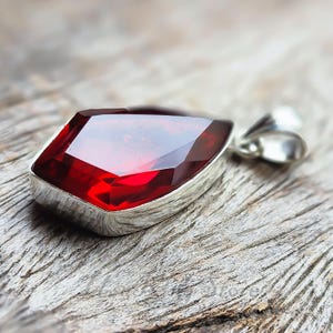 May include: A faceted, deep red gemstone pendant with a silver setting. The pendant has a geometric shape and a polished finish. The gemstone is set against a weathered wooden surface, highlighting its vibrant colour and intricate design.