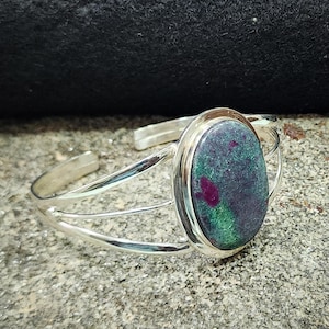 Puede incluir: Un brazalete de plata con una piedra ovalada verde y roja engastada en un bisel de plata. El brazalete tiene un diseño de triple banda.