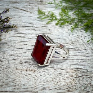 May include: A silver ring featuring a large, rectangular, deep red gemstone. The ring has a detailed band and a setting that frames the stone. The ring is displayed on a weathered wooden surface, with greenery in the background.