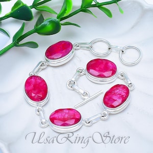 Puede incluir: Una pulsera de plata con piedras preciosas rojas brillantes, de forma ovalada y facetadas. Las piedras están engastadas en un bisel de plata y unidas por eslabones de plata. La pulsera se muestra sobre una superficie blanca con follaje verde.