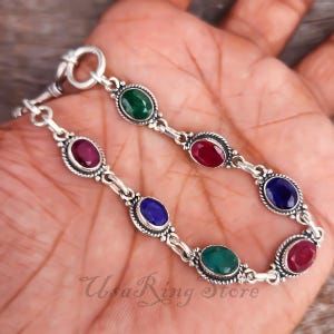 May include: Silver bracelet featuring oval-cut gemstones in red, green, and blue. The bracelet has a decorative border around each stone and a toggle clasp. The text "Usa Ring Store" is visible on the bracelet.