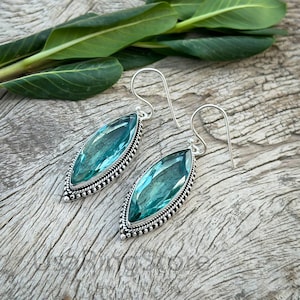 Puede incluir: Un par de pendientes de plata con una piedra preciosa azul verdosa en forma de lágrima engastada en un engaste de plata. Los pendientes tienen un borde de cuentas alrededor de la piedra preciosa.