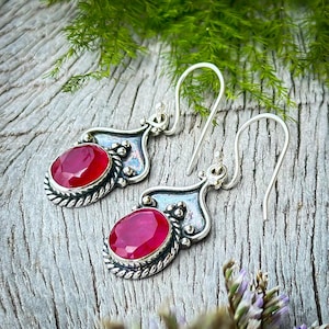 May include: A pair of silver earrings with red gemstone drops. The earrings have a delicate, ornate design with a twisted rope border around the gemstone.