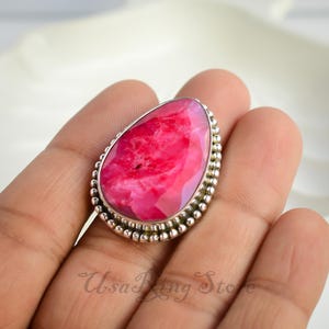 May include: A silver ring with a large, teardrop-shaped pink gemstone. The ring has a beaded edge.