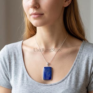 Puede incluir: Un colgante rectangular de lapislázuli de color azul intenso con motas naturales de color dorado, engastado en un marco de plata. El colgante cuelga de una fina cadena de plata sobre una camiseta gris. El texto UsaRingStore es visible.