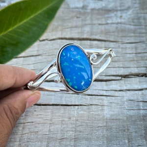 Peut inclure: Un bracelet manchette en argent avec une grande opale bleue ovale. L'opale est d'un bleu vif avec des éclats verts. Le bracelet a un design simple avec des détails argentés incurvés. Bijou élégant.