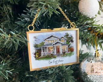 Puede incluir: Un adorno rectangular de metal dorado que cuelga de una cadena, con una ilustración estilo acuarela de una casa de dos pisos con porche. El texto "Our First Home" y "2026" está impreso en negro en la parte inferior de la imagen.