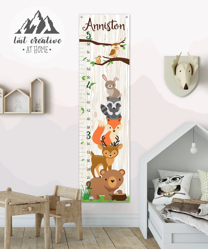 May include: A white wooden growth chart with a woodgrain pattern features a whimsical woodland theme. The chart is decorated with a brown bear, a deer, a fox, a raccoon, a rabbit, and an owl. The chart is labeled with numbers from 20 to 58 inches. The chart is personalized with the name "Anniston".
