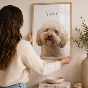 May include: A framed portrait of a fluffy, light brown dog with its tongue out. The dog's name, "Oliver," is written above the image in cursive. The frame is light wood, and the portrait is being held up.