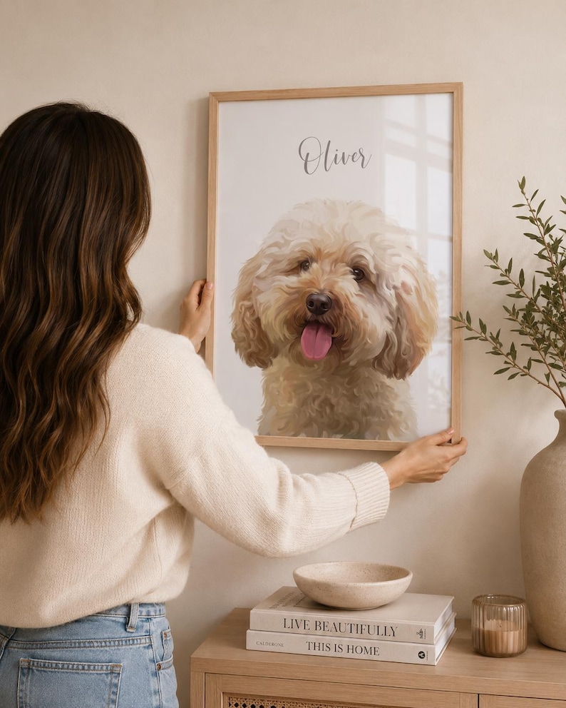 May include: A framed portrait of a fluffy, light brown dog with its tongue out. The dog's name, "Oliver," is written above the image in cursive. The frame is light wood, and the portrait is being held up.