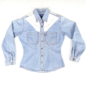 May include: Pale blue denim shirt with long sleeves, two chest pockets, and a button-down front. The shirt has white shoulder panels with embroidered floral designs. A red Levi's tag is visible on the left pocket.