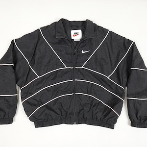 May include: Black Nike windbreaker jacket with white detailing. It has a full zip, elasticated cuffs, and a small Nike logo. The jacket's design is classic and sporty.