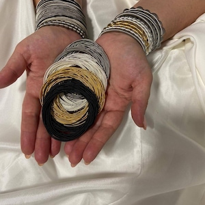 May include: A stack of gold, silver, and black bangle bracelets. The bracelets are arranged in a circle and are held in a person's hand.