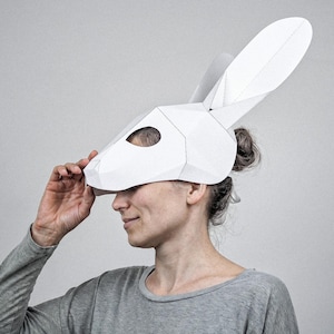 May include: A white, geometric rabbit mask with long ears, made from folded paper. A person wears the mask, paired with a grey long-sleeved top. The background is a plain, light grey.