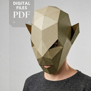 Puede incluir: Una máscara geométrica de papel en tonos beige y marrón, con orejas puntiagudas y un diseño angular detallado. La máscara la lleva una persona con una camiseta gris. Las palabras "DIGITAL FILES PDF" están en la esquina superior izquierda. El logotipo "simaga PAPER & WOOD" está en la parte inferior.