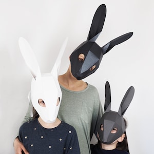 May include: Three geometric rabbit masks in white, black, and grey. The masks are worn by three people, with the white mask in the front. The masks have long, pointed ears and are made of folded paper or card. The background is a plain white wall.