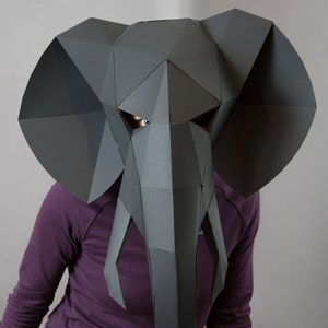 May include: A dark gray, low-poly geometric elephant mask constructed from paper panels. The mask features large, flat, angular ears, a central trunk, and two downward-pointing tusks, with cutouts for the eyes revealing a glimpse of the wearer.