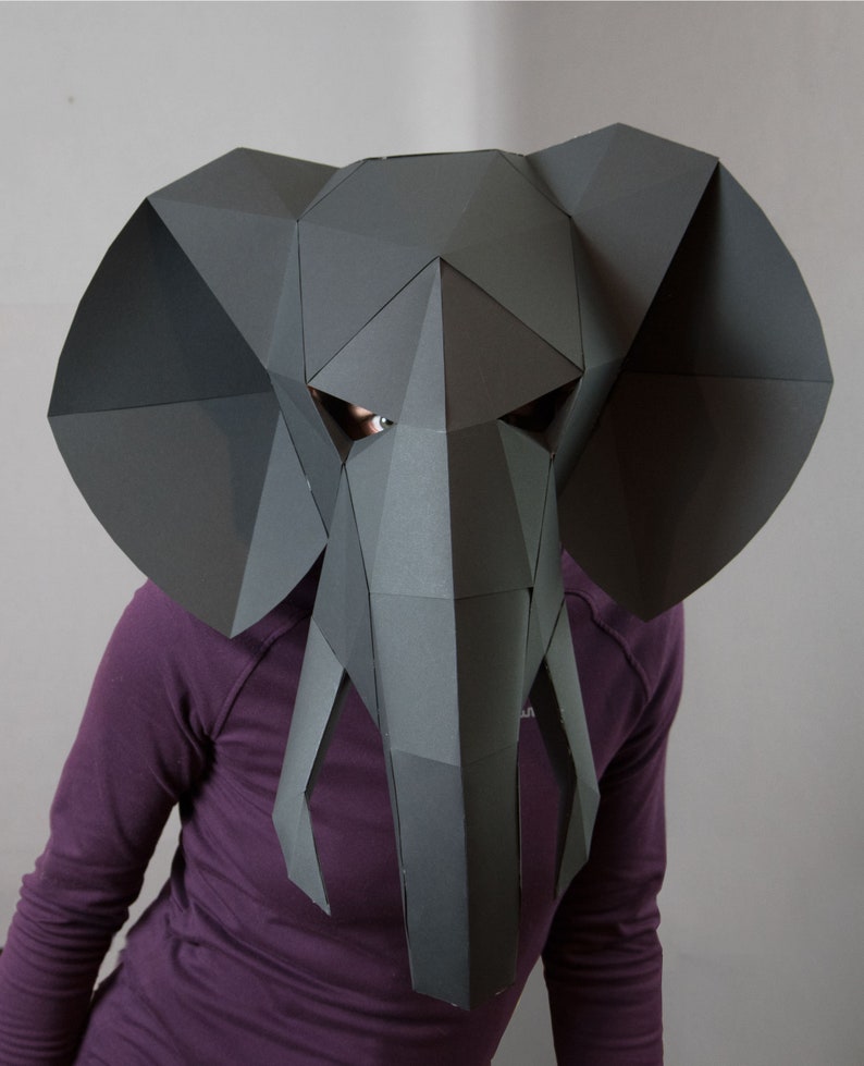 May include: A dark gray, low-poly geometric elephant mask constructed from paper panels. The mask features large, flat, angular ears, a central trunk, and two downward-pointing tusks, with cutouts for the eyes revealing a glimpse of the wearer.